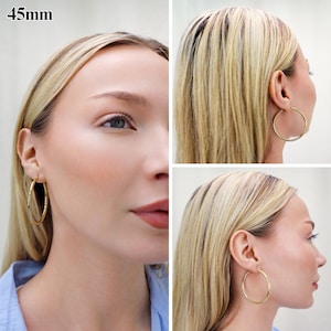 May include: A pair of gold-toned hoop earrings with a smooth, polished finish. The circular earrings have a diameter of 45mm. The hoops are shown from multiple angles, highlighting their simple, round, and continuous design worn in the earlobes.
