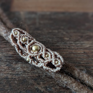 May include: A close-up of a brown and beige macrame hair wrap with gold beads. The wrap is tied around two brown dreadlocks.