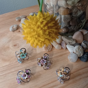May include: Four handmade wire rings with floral designs. Each ring features a different colored flower with rhinestones and gold accents. The rings are displayed on a wooden surface.