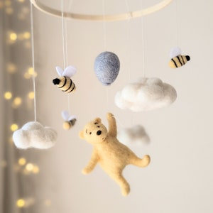 May include: A felt baby mobile with a yellow bear, white clouds, and bees. The bear is holding a blue balloon.