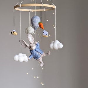 May include: A felt baby mobile with a blue bunny, clouds, a bee, a hot air balloon, and a carrot. The mobile is hanging from a wooden ring.