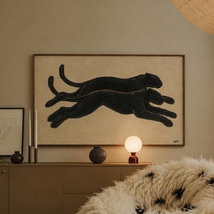 Puede incluir: Un puf grande, redondo y peludo cubierto de piel sintética larga de color crema con manchas negras dispersas. El sillón está situado en una habitación con una obra de arte mural horizontal enmarcada que representa dos panteras negras.