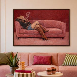 Op de afbeelding: Een groot rechthoekig ingelijst schilderij met een persoon in een hoed en laarzen die op een roze bank ligt terwijl diegene rookt. Het kunstwerk heeft een dieprode achtergrond en hangt boven een roze bank met diverse kussens en bijzettafels.