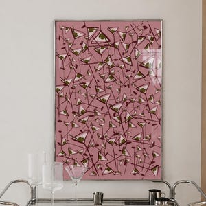 May include: A rectangular framed wall art piece featuring a repeating pattern of martini glasses in dark red and green on a dusty pink background, displayed above a two-tiered chrome bar cart with glass shelves holding various glassware and books.