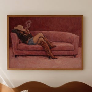 Może przedstawiać: Oprawiony obraz przedstawia osobę leżącą na różowej sofie, w kapeluszu kowbojskim i butach, palącą papierosa. Dzieło sztuki wykorzystuje ciepłą paletę barw, z odcieniami różu i brązu. Obraz jest zamontowany na jasnej ścianie.