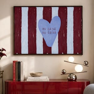 May include: A rectangular wall art piece featuring vertical maroon and light blue stripes with a central light blue heart shape containing the text 'mi casa su casa' in dark red, framed in a thin dark brown border hanging above a red sideboard.