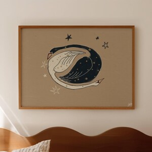 Op de afbeelding: Een ingelijste rechthoekige wandkunstprint met een donkerblauwe en crèmekleurige illustratie van twee zwanen die in een cirkelvorm in elkaar verstrengeld zijn, omringd door kleine witte sterren op een beige achtergrond, in een dunne houten lijst.