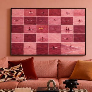 Könnte beinhalten: Ein rechteckiges Wandbild mit einem Raster aus rosa und dunkelroten Quadraten, die jeweils kleine gemalte Figuren beim Schwimmen oder Surfen zeigen, inklusive eines gelben Schwimmrings, gerahmt in einem dünnen schwarzen Rahmen über einem Sofa.