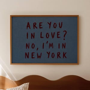 May include: A rectangular wall art print in a thin wooden frame featuring a dark blue background with the text in dark red, hand-drawn style lettering: ARE YOU IN LOVE? NO, I'M IN NEW YORK. The print is displayed horizontally on a wall.