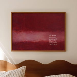 May include: A framed art print featuring a deep red abstract design and the text "12 YEAR OLD YOU WOULDN'T BELIEVE HOW COOL YOU ARE." The artwork is displayed above a bed with striped pillows and a wooden headboard.