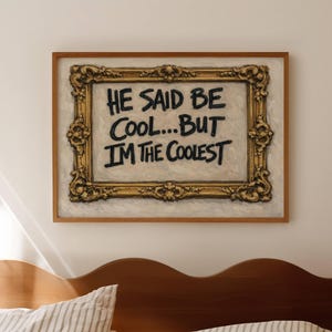 May include: A rectangular wall art piece featuring a gold ornate frame surrounding a white canvas with the black hand-lettered text: HE SAID BE COOL...BUT I'M THE COOLEST. The artwork is displayed in a simple wooden frame border.