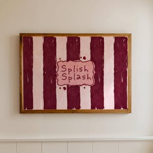 May include: A framed artwork with a wooden frame hangs on a wall. The artwork features vertical stripes in burgundy and light pink. In the center, the words "Splish Splash" are written in a playful font within a pink cloud-shaped design.