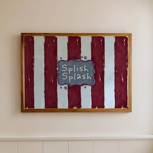 May include: A framed artwork with a wooden frame. The artwork features vertical stripes in burgundy and light blue. The words "Splish Splash" are written in a blue cloud-shaped text box. The artwork is hanging on a cream-colored wall.