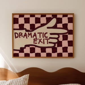 May include: A rectangular framed wall art piece featuring a cream-coloured hand pointing to the right against a dark maroon and light pink checkered background. The text "DRAMATIC EXIT" is printed in dark maroon letters across the hand graphic.