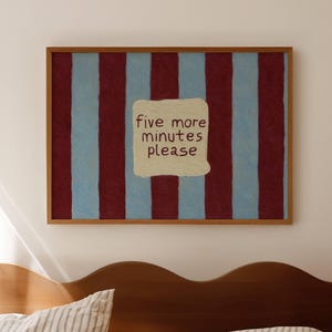Può includere: Un'opera d'arte da parete rettangolare in una cornice di legno con strisce verticali che si alternano tra il rosso scuro e il blu chiaro. Al centro delle strisce si trova una toppa color crema con il testo scritto a mano five more minutes please.