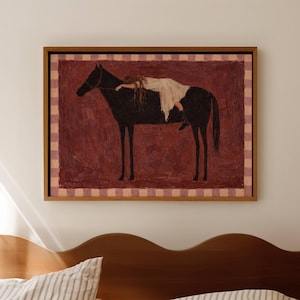 May include: A framed wall art print featuring a dark brown horse standing in profile against a textured deep red background. A figure in a white dress lies across the horse's back. The artwork is bordered by a light pink and white checkered frame.