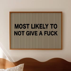 Puede incluir: Un cuadro enmarcado con el texto "MOST LIKELY TO NOT GIVE A FUCK" en letras negras. El fondo tiene rayas verticales beige y blancas. El marco es de color marrón. La obra de arte cuelga sobre una cama.