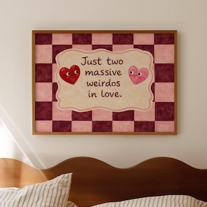 May include: A framed print with the text "Just two massive weirdos in love." The artwork features a pink and burgundy checkerboard background with a central cream-colored shape. Two cartoon hearts, one red and one pink, are on the artwork.