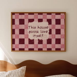 May include: A framed artwork with a checkered pattern in shades of pink and burgundy. The artwork features the text "This house gonna love itself" in a handwritten style. The frame is made of wood, and the artwork is hanging above a bed.