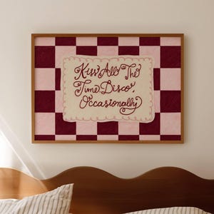 Puede incluir: Una obra de arte enmarcada con un fondo de tablero de ajedrez en tonos rosa y burdeos. La obra presenta un rectángulo crema con bordes festoneados con el texto manuscrito "Kiss All The Time Disco Occasionally" en escritura burdeos.
