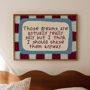 May include: A framed art print with the text "Those dreams are actually really silly but I think I should chase them anyway." The artwork has a light blue border and a red and white checkered background. The frame is brown.