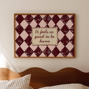 May include: A framed print with a pink and burgundy diamond pattern. The artwork features the text "It feels so good to be home" in a decorative font. The frame is a warm brown colour, and the artwork is displayed above a bed.
