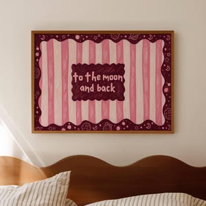 May include: A framed artwork with a whimsical design. The artwork features vertical pink and white stripes, a dark red border with celestial motifs, and a central dark red rectangle with the text "to the moon and back". The frame is a warm brown color.