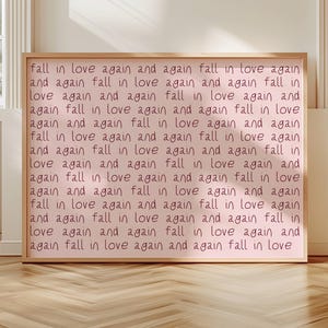 May include: A rectangular framed wall art piece featuring a light pink background with the repeating dark pink handwritten text "fall in love again and again" covering the entire surface in a grid-like pattern, enclosed in a light wood frame.