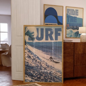 May include: Three framed art prints with a surfing theme. The largest print features the word "SURF" in large blue letters above a surfer on a wave. Two smaller prints display abstract designs and the text "RIDE THE WAVE".