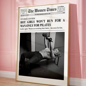May include: A framed black and white print of a newspaper front page titled "The Women Times." The headline reads "Hot Girls Won't Run For A Man-Only For Pilates." The image features a woman lifting weights. The frame is gold, and the background is pink.