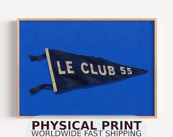 May include: A framed wall art print featuring a dark navy blue triangular pennant flag centered on a solid bright blue background. The pennant displays the white text LE CLUB 55 and includes two black fabric ties on the left side attached to a light wooden pole.