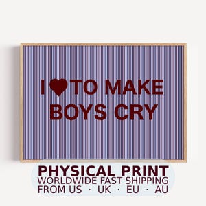 Puede incluir: Una lámina decorativa rectangular en un marco de madera clara con un patrón de rayas verticales en azul oscuro y morado claro. El texto I (símbolo de corazón) TO MAKE BOYS CRY está impreso en una fuente negrita de color rojo oscuro.
