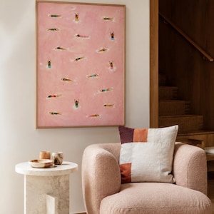 May include: A framed rectangular wall art piece featuring a pink background with numerous small painted figures of people swimming in various positions. The artwork is displayed above a textured pink armchair with a multi-colored square throw pillow.