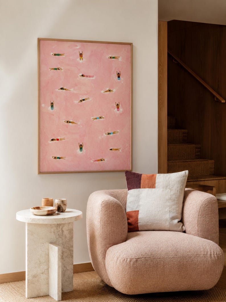 May include: A framed rectangular wall art piece featuring a pink background with numerous small painted figures of people swimming in various positions. The artwork is displayed above a textured pink armchair with a multi-colored square throw pillow.