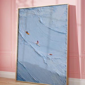 Puede incluir: Una pieza de arte mural rectangular enmarcada que presenta una pintura al óleo con técnica de empaste, gruesa y texturizada, de una superficie oceánica azul claro. Tres pequeñas figuras son visibles en el agua: una en una tabla de surf naranja, una en una tabla de surf blanca y una en un kayak rojo.
