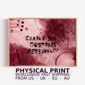 May include: A rectangular wall art print in a light wooden frame featuring an abstract background of various shades of deep red, burgundy, and soft pink. The text "COMPARISON DESTROYS PERSONALITY" is printed in bold, black, hand-lettered style font.