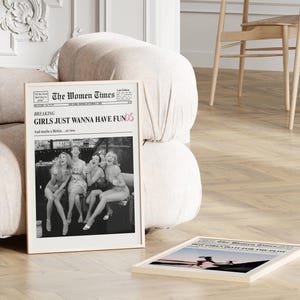 Op de afbeelding: Een ingelijste verticale poster getiteld The Women Times met een zwart-witfoto van vier lachende mensen. De kop luidt Breaking Girls Just Wanna Have Funds en And maybe a Birkin... or two. Een tweede ingelijste poster ligt op de grond.