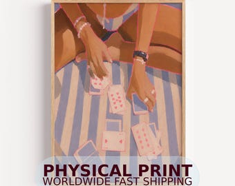 May include: A framed art print depicting a pair of hands playing with a deck of cards on a blue and white striped surface. The hands are adorned with bracelets and rings, and the cards feature pink heart symbols. The artwork is presented in a light wood frame.