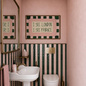Puede incluir: Un ba&ntilde;o con paredes rosas y azulejos a rayas verticales en verde oscuro y rosa. Un lavabo de pedestal blanco con grifo de lat&oacute;n, un inodoro suspendido, un espejo con marco de lat&oacute;n y un cuadro con el texto I SEE LONDON I SEE FRANCE.