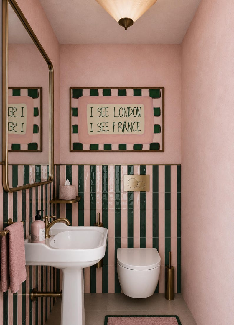 Puede incluir: Un ba&ntilde;o con paredes rosas y azulejos a rayas verticales en verde oscuro y rosa. Un lavabo de pedestal blanco con grifo de lat&oacute;n, un inodoro suspendido, un espejo con marco de lat&oacute;n y un cuadro con el texto I SEE LONDON I SEE FRANCE.