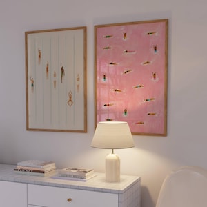 May include: Two framed rectangular wall art prints featuring illustrations of swimmers in a pool. The left print has a white background with vertical blue lines, while the right print has a pink background with multiple swimmers scattered throughout.