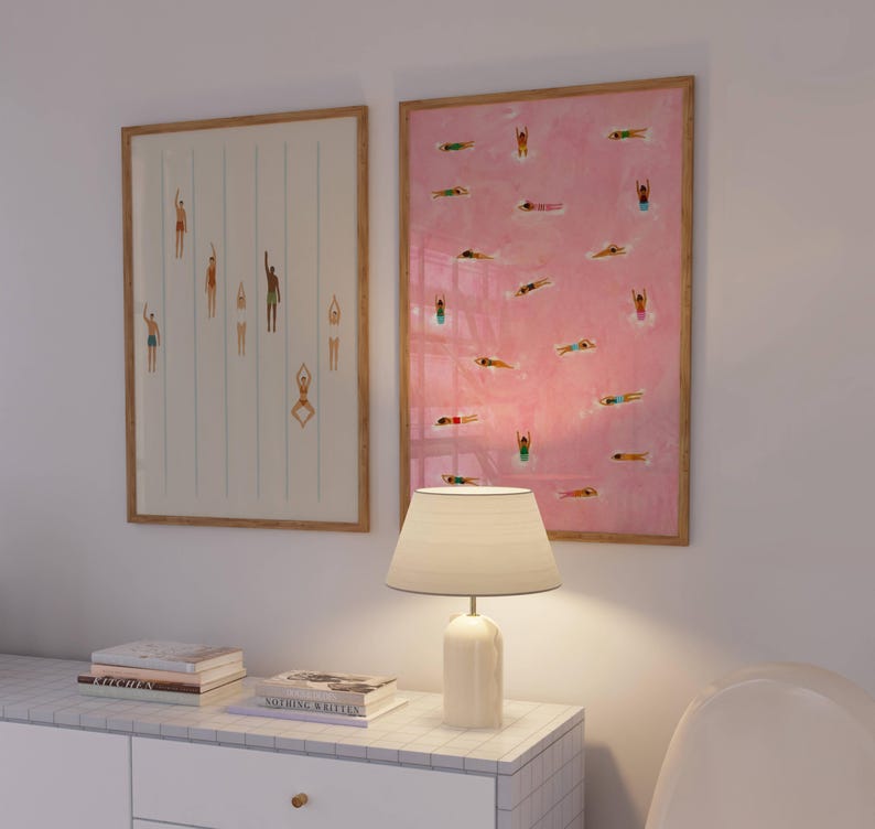 May include: Two framed rectangular wall art prints featuring illustrations of swimmers in a pool. The left print has a white background with vertical blue lines, while the right print has a pink background with multiple swimmers scattered throughout.