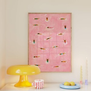 May include: A framed rectangular wall art print featuring a pink background with numerous small figures of people swimming in various colorful swimsuits. The artwork is displayed alongside a yellow mushroom-shaped lamp and two candles.