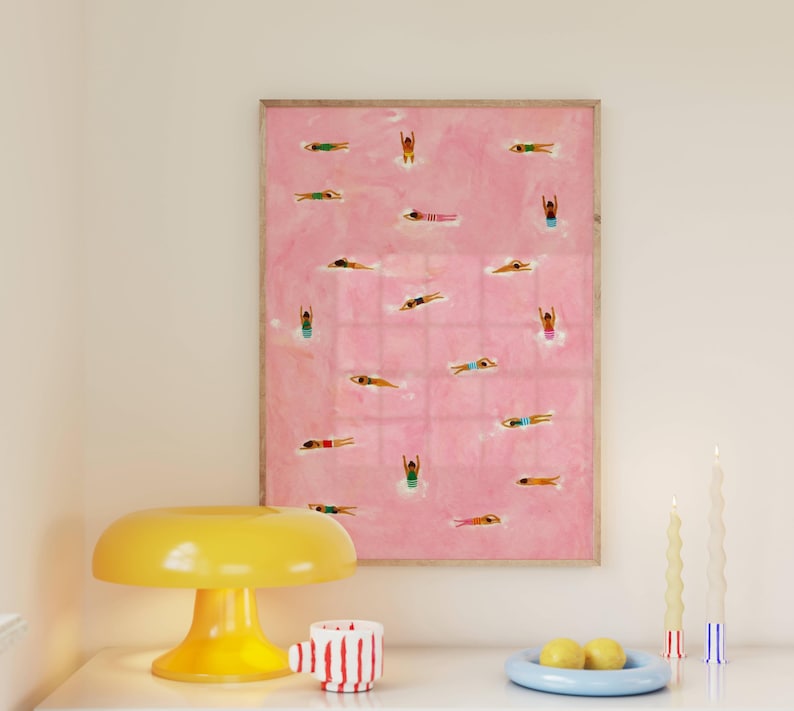 May include: A framed rectangular wall art print featuring a pink background with numerous small figures of people swimming in various colorful swimsuits. The artwork is displayed alongside a yellow mushroom-shaped lamp and two candles.