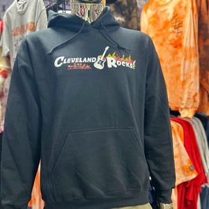 Op de afbeelding: Zwarte hoodie met een voorzak en een capuchon met trekkoord. De hoodie toont de tekst "Cleveland Rocks" in het wit met een gitaar- en vlammenafbeelding. Andere kleding is zichtbaar op de achtergrond.