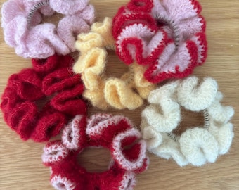 May include: A collection of six handmade scrunchies in various colors. The scrunchies are made of a soft, textured yarn and include shades of pink, red, yellow, and cream. Each scrunchie is approximately 4 inches in diameter.