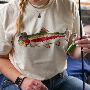 Pode incluir: Uma camiseta branca com uma truta colorida usando um chapéu impressa na frente. A truta está virada para a direita e tem uma faixa vermelha no corpo. A pessoa que está usando a camiseta está segurando uma vara de pesca.