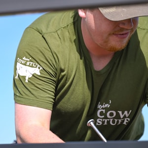 May include: A man wearing a green t-shirt with the text "doin' COW STUFF" and a white cow graphic on the left sleeve. The text "doin' COW STUFF" is also on the right side of the shirt.