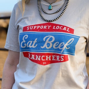 May include: A white t-shirt with a red, white, and blue graphic that says "Support Local Eat Beef Ranchers" with a small "XM" at the bottom.