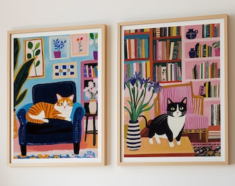 May include: Two framed art prints featuring colorful illustrations of cats in cozy settings. One print shows an orange cat on a blue armchair, the other a black and white cat in a room with bookshelves. Both are in light wood frames.